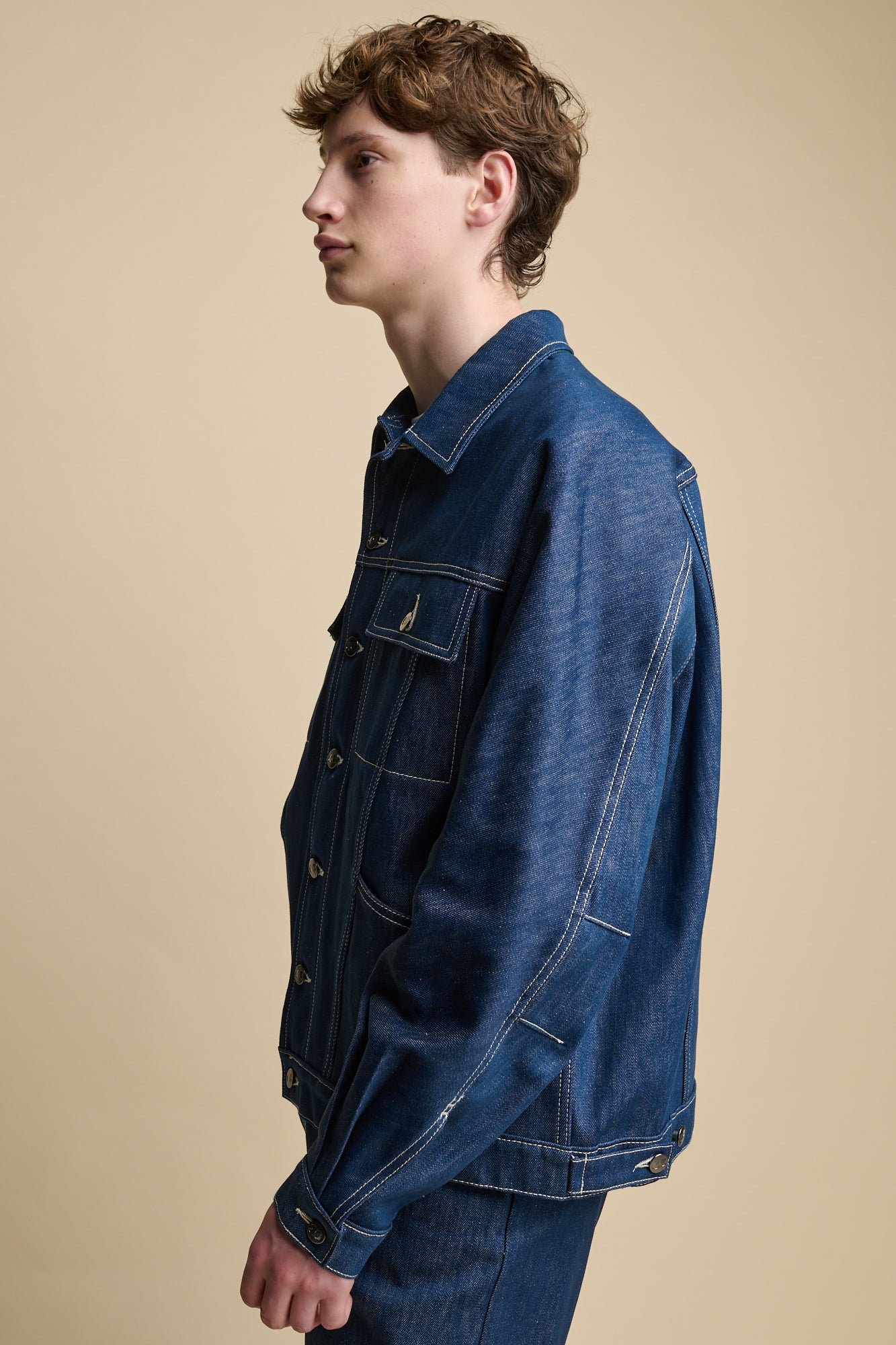 Portrait of white male wearing Joe Denim Jacket in blue over short sleeve t shirt in white. Jacket has contrast white stitching and silver branded buttons, two front pockets with flaps