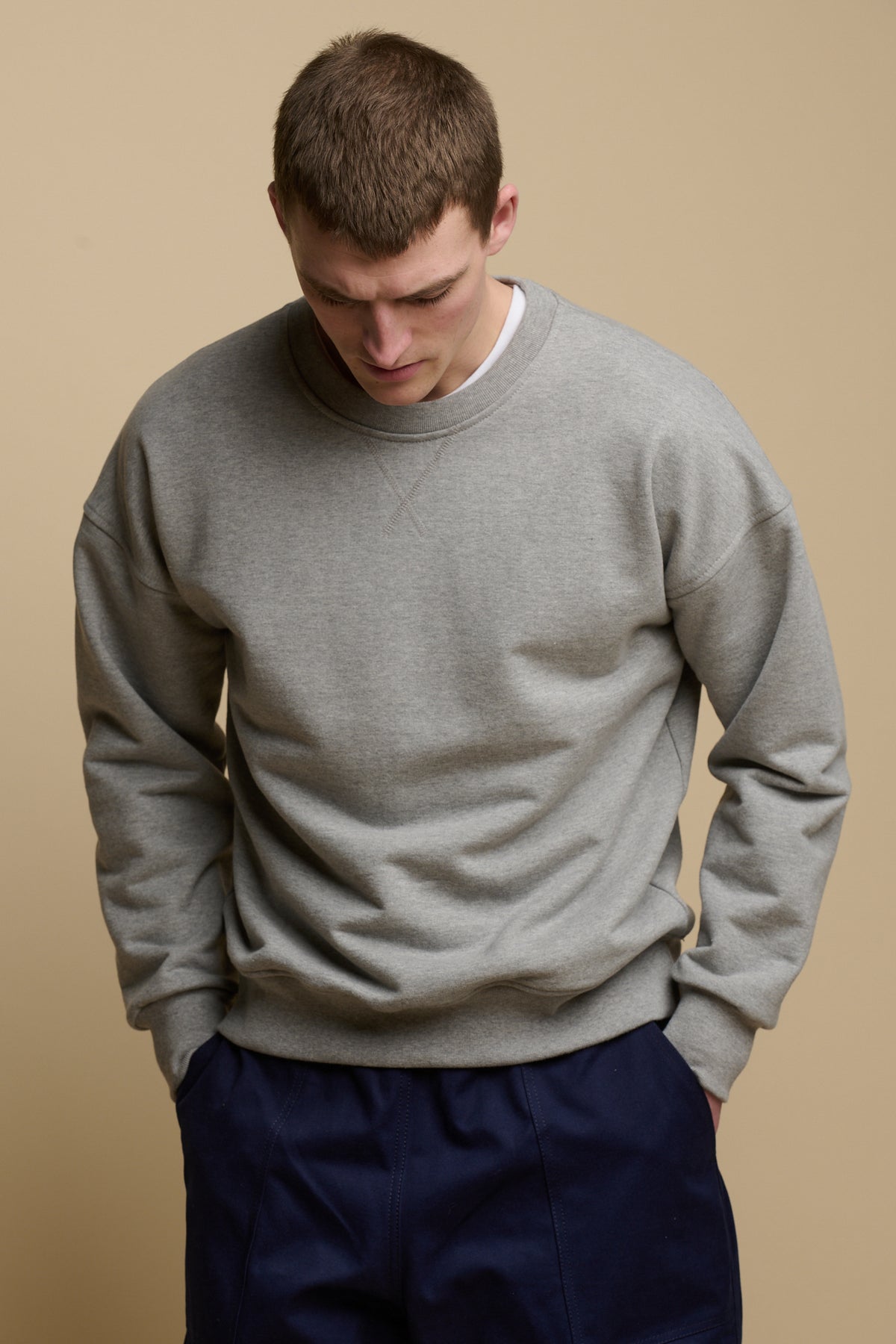 
            Thigh up front of brunet male looking down to the ground, wearing men&#39;s heritage drop shoulder sweatshirt in grey, worn over crew neck white t shirt paired with cameraman pants in navy